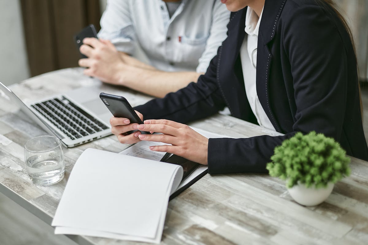 Homem e mulher com celular em mãos, empresários, conversando em frente a um notebook sobre: Quais são as vantagens de uma gestão de assinatura digital?